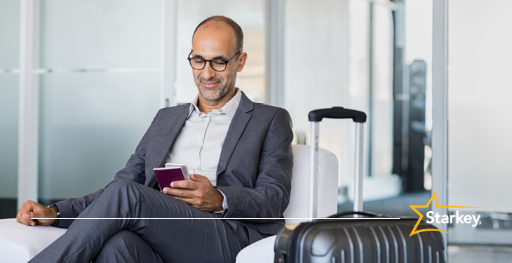 man in grey suit sitting at an airport and looking at his phone with a carry-on next to him 